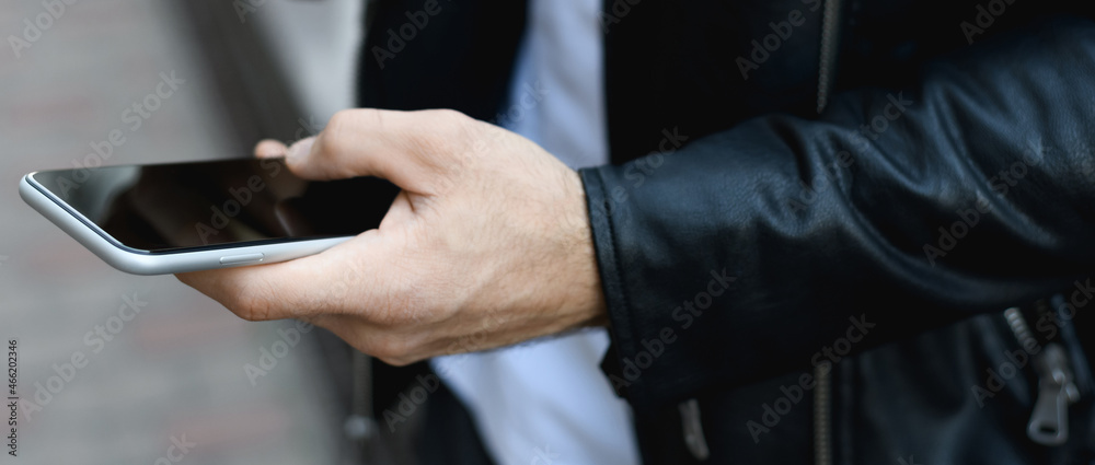 Cropped view of man in leather jacket holding smartphone with blank screen outdoors, banner
