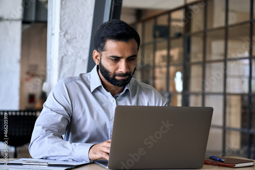 Handsome serious concentrated focused young Indian Hispanic ceo businessman using computer working in modern office, having virtual training on laptop typing watching online webinar.