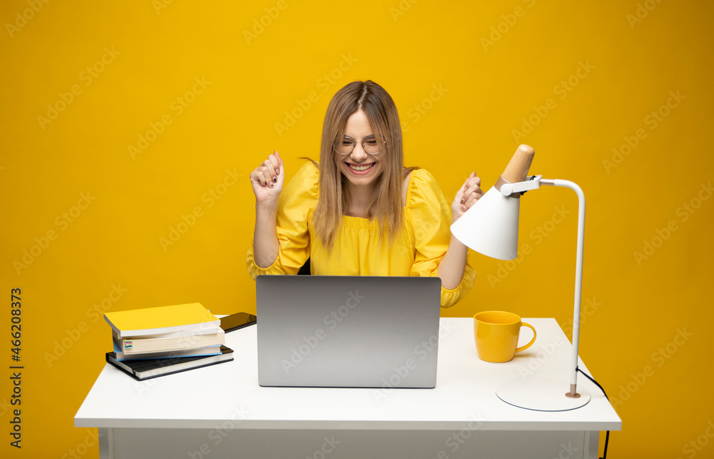 Excited young woman working with a grey laptop computer, notebook while sitting at the table with grey laptop. Smiling business woman or student received a good news isolated on a yellow background. Excited young woman working with a grey laptop computer, notebook while sitting at the table with grey laptop. Smiling business woman or student received a good news isolated on a yellow background.