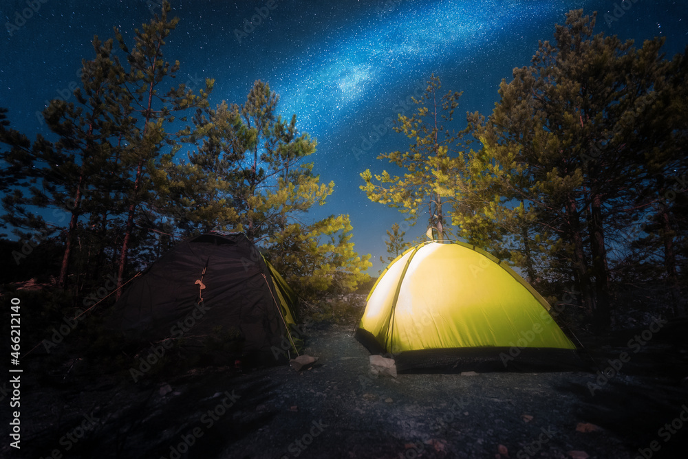 Small yellow tent with illumination inside in the middle of the forest at night with starry sky. Camping under the Milky Way. Tourism.