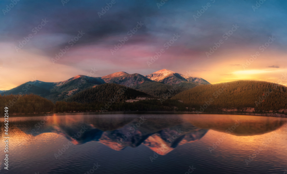 Beautiful landscape with mountains near the lake with reflection on water against blue cloudy sky and yellow sunlight at sunset.