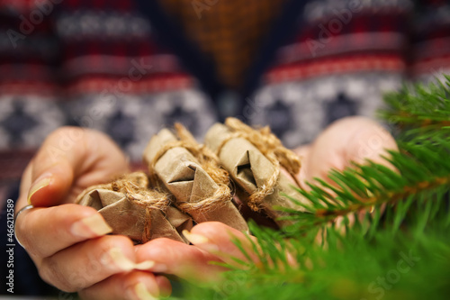 Close-up female hands with a three little craft gifts. Young hipster woman in Christmas sweater holding 3 organic gifts, rope bow. Cozy warm winter detail. Green natural foreground