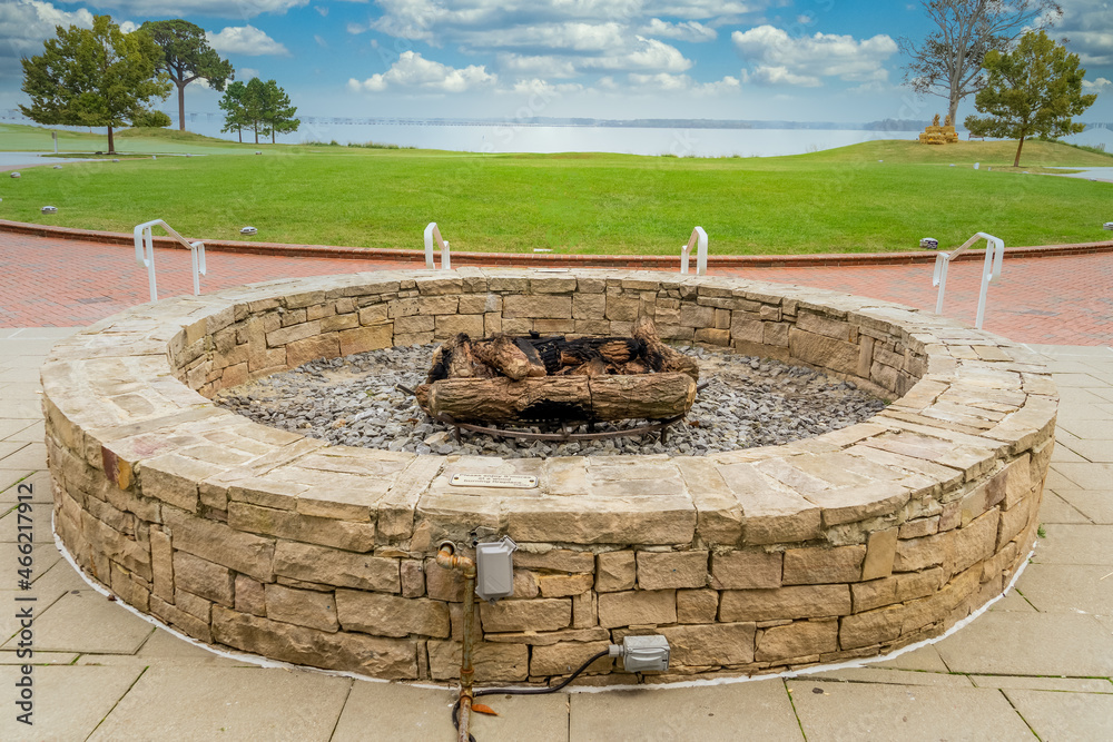 Outdoor patio gas fueled fireplace or firepit covered in stone veneer to roast marshmallows on the side of a lake and green grass