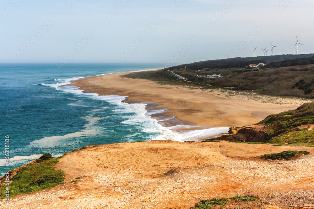 A beautiful landscape of the burgundy Atlantic Ocean and the sandy yellow shore with white foam from the waves. In the background are several houses and wind turbines of energy. Portugal, Nazare.