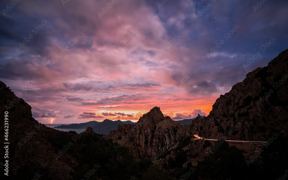 Sunrise over the Calanches of Piana in Corsica with lightning in the distance and car lights passing along the D81 road