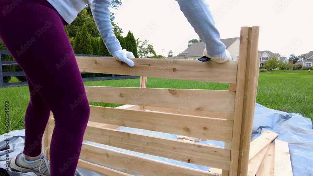a woman is building a wooden fence or keep on tarp in her backyard. A concept for wood work. Furniture installation DIY project. She wears gloves to protect against wood splinters