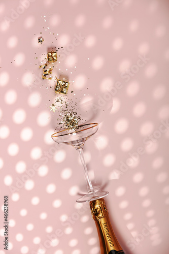 Bottle of champagne, glass with splash of confetti and golden decorations over pink background © Natalia Klenova