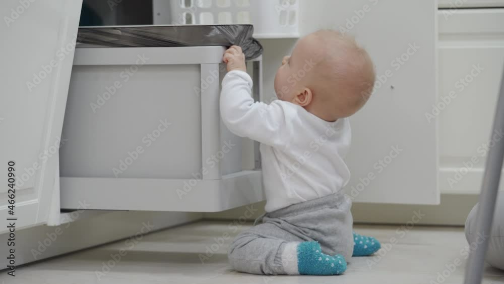 Home has many potential hazards for a child, baby opening cabinet grabbing and playing with pull out trash can under kitchen counter, safety at home for kids. High quality 4k footage Home has many potential hazards for a child, baby opening cabinet grabbing and playing with pull out trash can under kitchen counter, safety at home for kids. High quality 4k footage