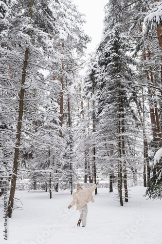 Happy girl in warm clothes spinning around on snowy ground in winter forest. Trees covered by snow. Happiness concept