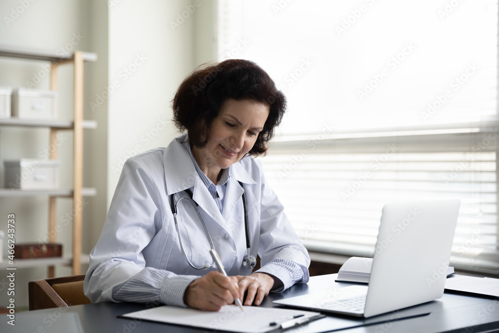 Smiling middle aged female doctor general practitioner making notes on paper in clipboard, making notes in disease history, writing prescription for medication, working alone in clinic office. Smiling middle aged female doctor general practitioner making notes on paper in clipboard, making notes in disease history, writing prescription for medication, working alone in clinic office.