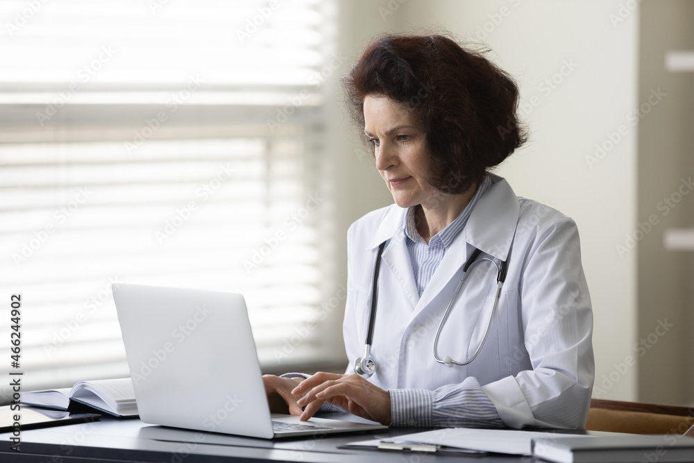 Concentrated middle aged female doctor physician in white coat works on computer, consulting patients distantly, web surfing medical information, prescribing illness treatment online at clinic office. Concentrated middle aged female doctor physician in white coat works on computer, consulting patients distantly, web surfing medical information, prescribing illness treatment online at clinic office.