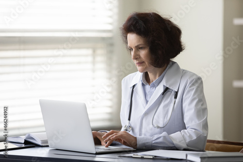 Concentrated middle aged female doctor physician in white coat works on computer, consulting patients distantly, web surfing medical information, prescribing illness treatment online at clinic office.