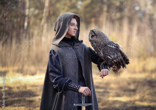 Fotografie Male warrior in grey cloak and long sword in hand with owl on his shoulder standing in the forest