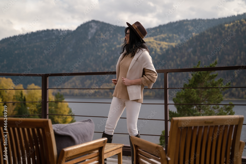 beautiful girl in stylish light-colored clothes with sunglasses brown hat stands on roof building rays sun against the backdrop of mountains lake. Autumn yellow trees and chill. High quality photo