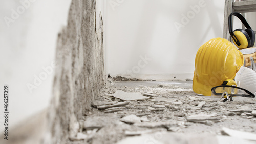 house renovation concept, wall in demolition with plaster rubble and protective construction work tools on the ladder, yellow helmet, glasses and headphones on white background with copy space
