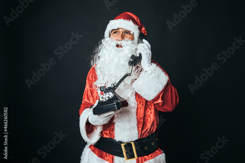 Man dressed as Santa Claus holding a retro telephone from the 60s, talking, on black background. Concept of Christmas, Santa Claus, gifts, celebration.