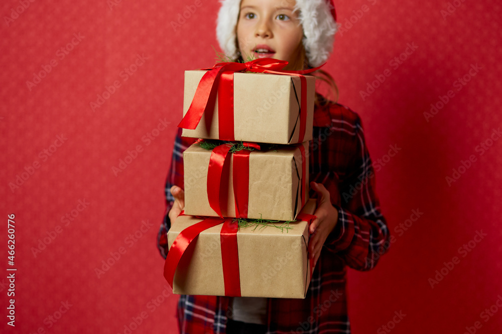 Funny little girl in santa hat with many Christmas gift boxes isolated on red background,