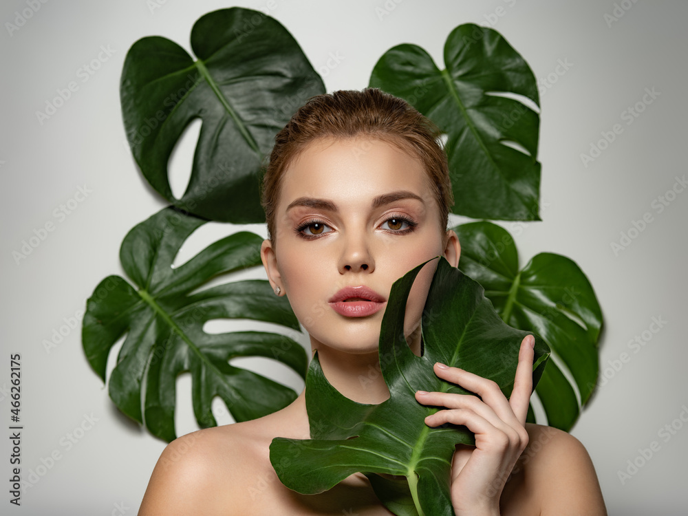 Closeup face of young beautiful woman with a healthy clean skin. Beautiful white girl with big green leaves. Beauty and spa treatment concept. Pretty woman with natural makeup and plant near face Closeup face of young beautiful woman with a healthy clean skin. Beautiful white girl with big green leaves. Beauty and spa treatment concept. Pretty woman with natural makeup and plant near face