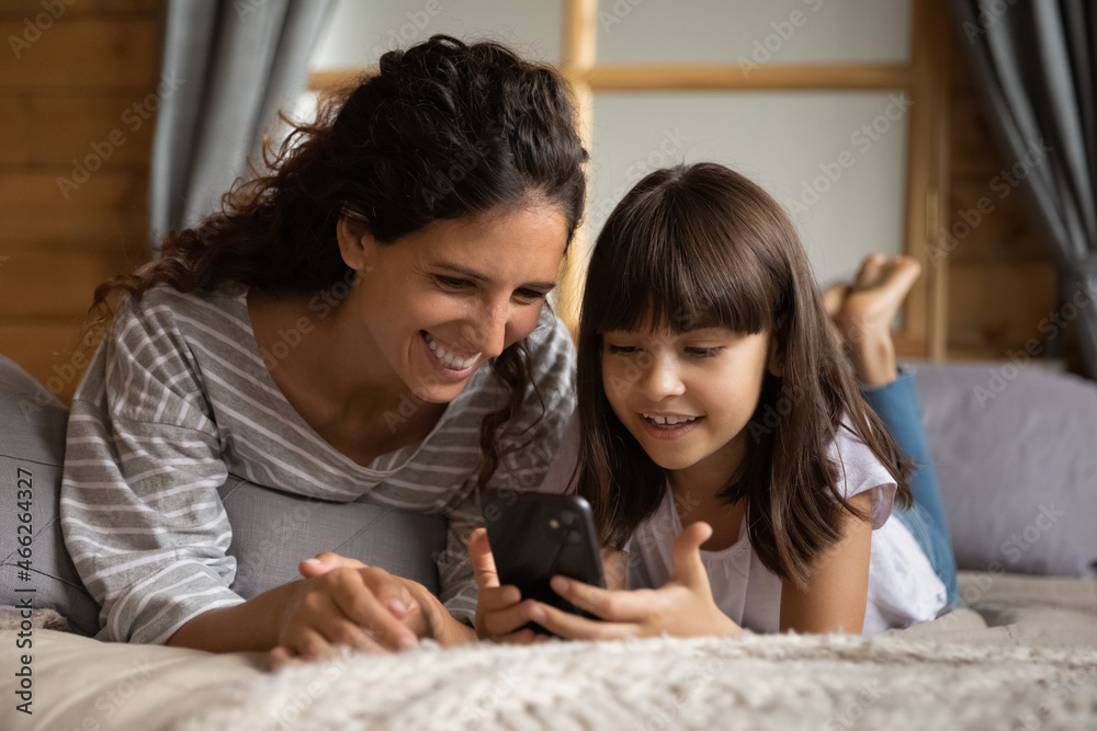 Happy young mom and daughter kid relaxing on bed, holding smartphone, using online app, virtual service, playing game, making call, reading book, watching media content, shopping on internet Happy young mom and daughter kid relaxing on bed, holding smartphone, using online app, virtual service, playing game, making call, reading book, watching media content, shopping on internet