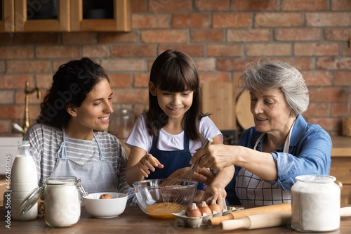 Happy Latin mom and grandma teaching girl to bake in kitchen. Mother  grandmother  kid cooking together  preparing dough for bakery food  mixing egg  flour in bawl on table. Family home activities