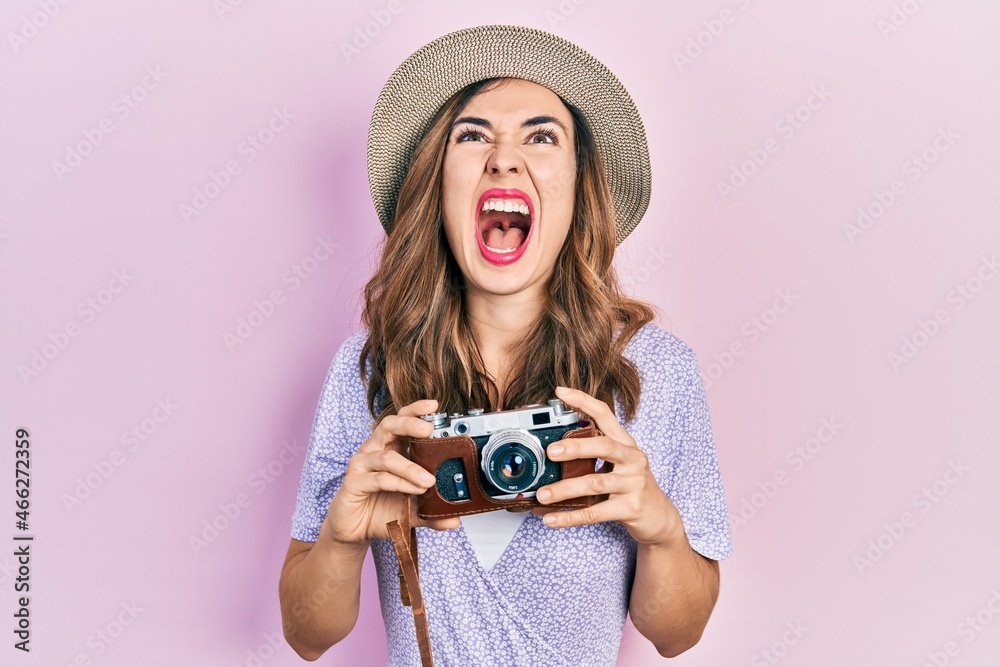 Young hispanic girl wearing summer hat holding vintage camera angry and mad screaming frustrated and furious, shouting with anger looking up.