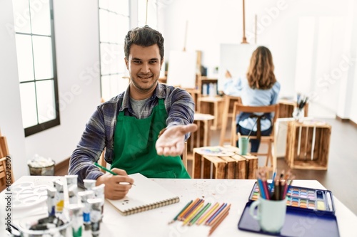 Young artist man at art studio smiling cheerful offering palm hand giving assistance and acceptance.