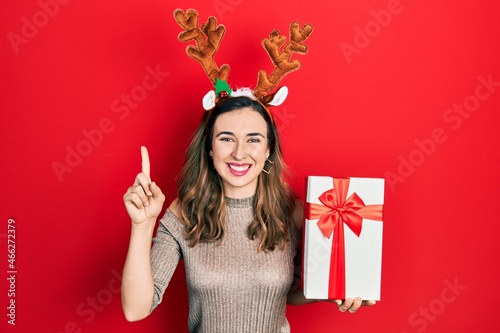 Young hispanic girl wearing deer christmas hat holding gift smiling with an idea or question pointing finger with happy face, number one