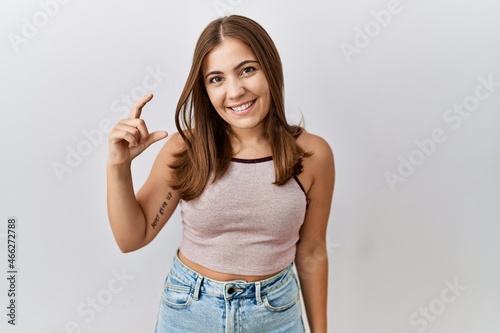 Young brunette woman standing over isolated background smiling and confident gesturing with hand doing small size sign with fingers looking and the camera. measure concept.