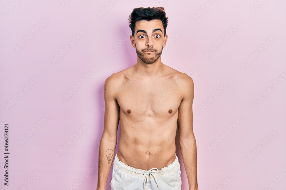 Young hispanic man wearing swimwear shirtless puffing cheeks with funny face. mouth inflated with air, crazy expression.