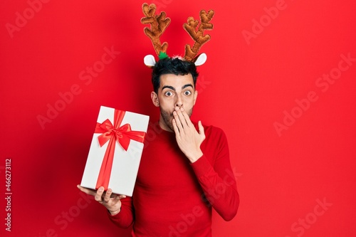 Young hispanic man wearing cute christmas reindeer horns holding gifts covering mouth with hand, shocked and afraid for mistake. surprised expression