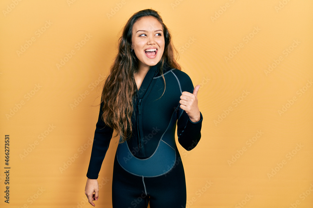 Young hispanic girl wearing diver neoprene uniform smiling with happy face looking and pointing to the side with thumb up. Young hispanic girl wearing diver neoprene uniform smiling with happy face looking and pointing to the side with thumb up.