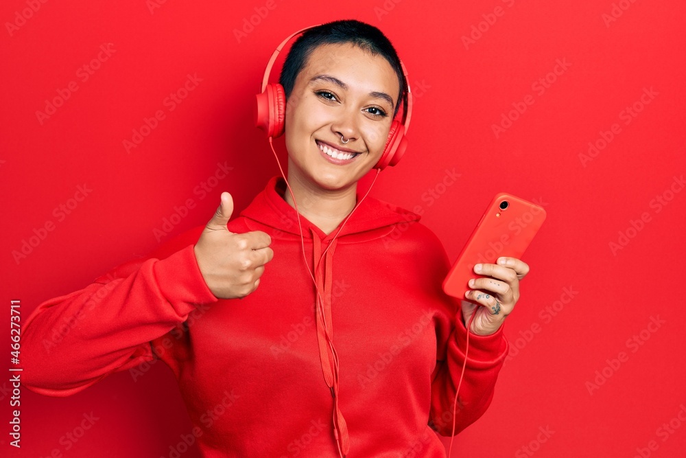 Naklejka premium Beautiful hispanic woman with short hair using smartphone wearing headphones smiling happy and positive, thumb up doing excellent and approval sign
