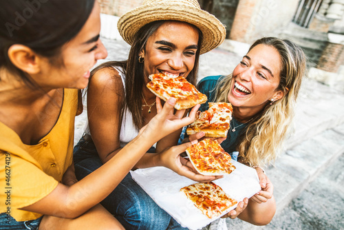 Three young female friends sitting outdoor and eating pizza - Happy women having fun enjoying a day out on city street - Happy lifestyle and tourism concept