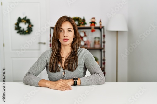 Young latin woman sitting on the table by christmas decor relaxed with serious expression on face. simple and natural looking at the camera.