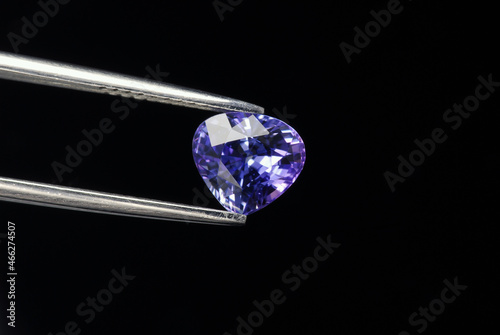 Natural purplish blue sapphire precious stone setting. Genuine, earth mined, clean, heart shaped, faceted loose gemstone specimen in tweezers. Black background. Gemology mineralogy jewelry theme.