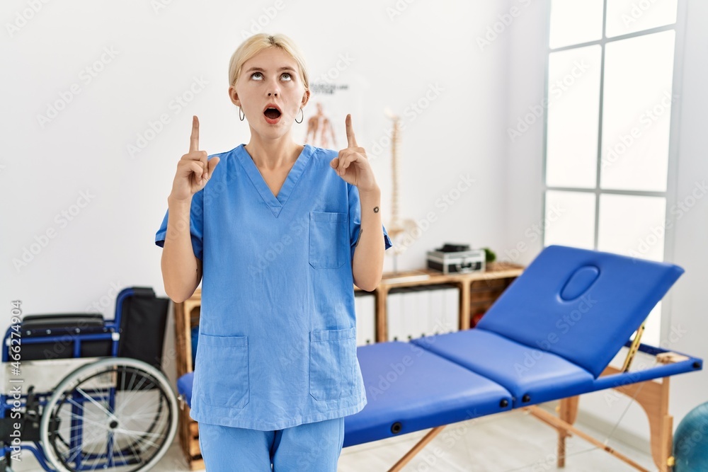 Beautiful caucasian physiotherapist woman working at pain recovery clinic amazed and surprised looking up and pointing with fingers and raised arms. Beautiful caucasian physiotherapist woman working at pain recovery clinic amazed and surprised looking up and pointing with fingers and raised arms.