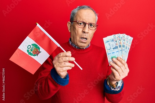 Handsome senior man with grey hair holding peru flag and peruvian sol banknotes clueless and confused expression. doubt concept.