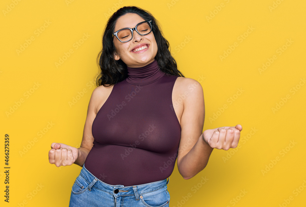Brunette young woman wearing casual clothes and glasses very happy and excited doing winner gesture with arms raised, smiling and screaming for success. celebration concept.