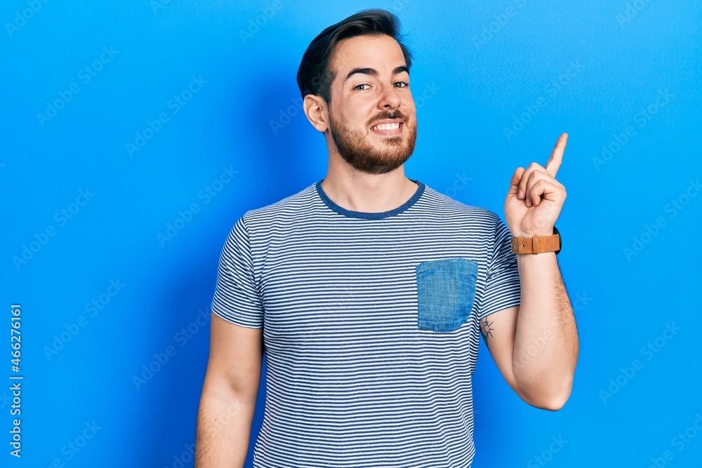 Handsome caucasian man with beard wearing casual striped t shirt with a big smile on face, pointing with hand and finger to the side looking at the camera. Handsome caucasian man with beard wearing casual striped t shirt with a big smile on face, pointing with hand and finger to the side looking at the camera.