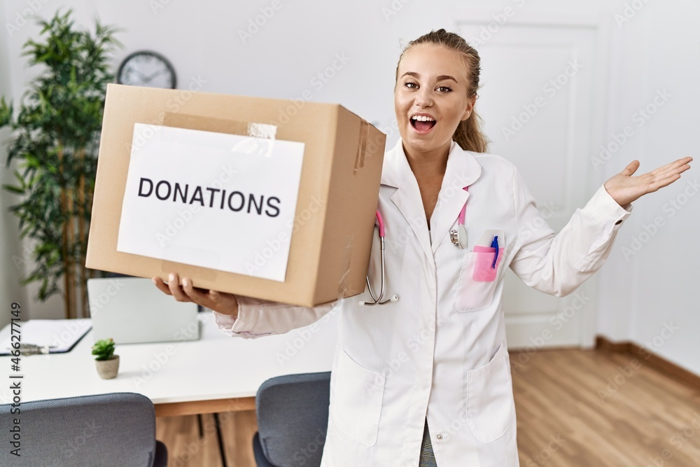 Young doctor woman at the clinic holding donations box celebrating achievement with happy smile and winner expression with raised hand Young doctor woman at the clinic holding donations box celebrating achievement with happy smile and winner expression with raised hand