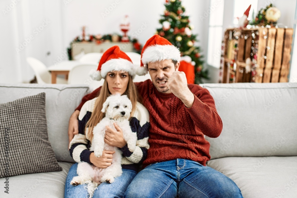 Obraz premium Young couple of wife and husband sitting on the sofa wearing christmas hat at home annoyed and frustrated shouting with anger, yelling crazy with anger and hand raised