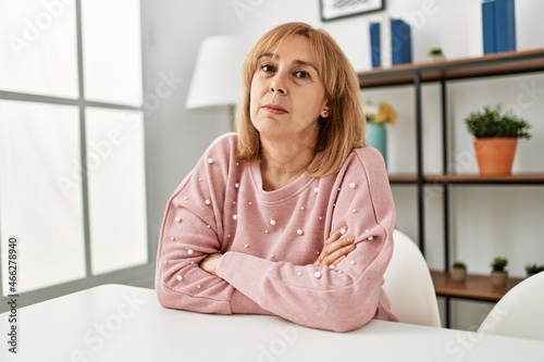 Middle age caucasian woman wearing casual clothes sitting on the table at home skeptic and nervous  disapproving expression on face with crossed arms. negative person.