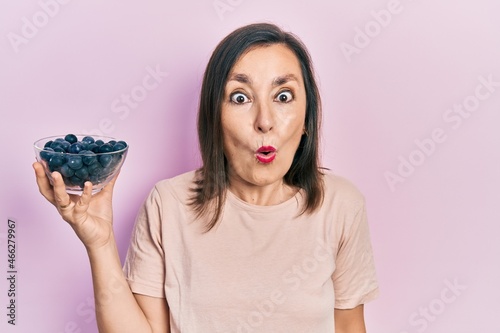 Middle age hispanic woman holding blueberries scared and amazed with open mouth for surprise, disbelief face