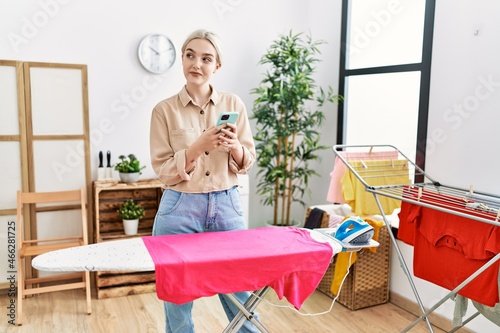 Young caucasian woman using smartphone ironing clothes at laundry room