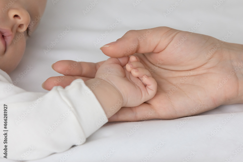 Crop close up loving mother touching small baby hand, sleeping on white bed, family enjoying tender moment, caring young mom holding cute infant newborn arm, motherhood and tenderness concept Crop close up loving mother touching small baby hand, sleeping on white bed, family enjoying tender moment, caring young mom holding cute infant newborn arm, motherhood and tenderness concept