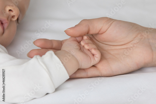 Crop close up loving mother touching small baby hand, sleeping on white bed, family enjoying tender moment, caring young mom holding cute infant newborn arm, motherhood and tenderness concept