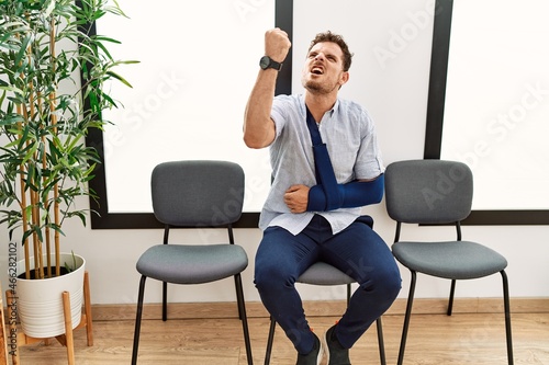 Handsome young man sitting at doctor waiting room with arm injury angry and mad raising fist frustrated and furious while shouting with anger. rage and aggressive concept.