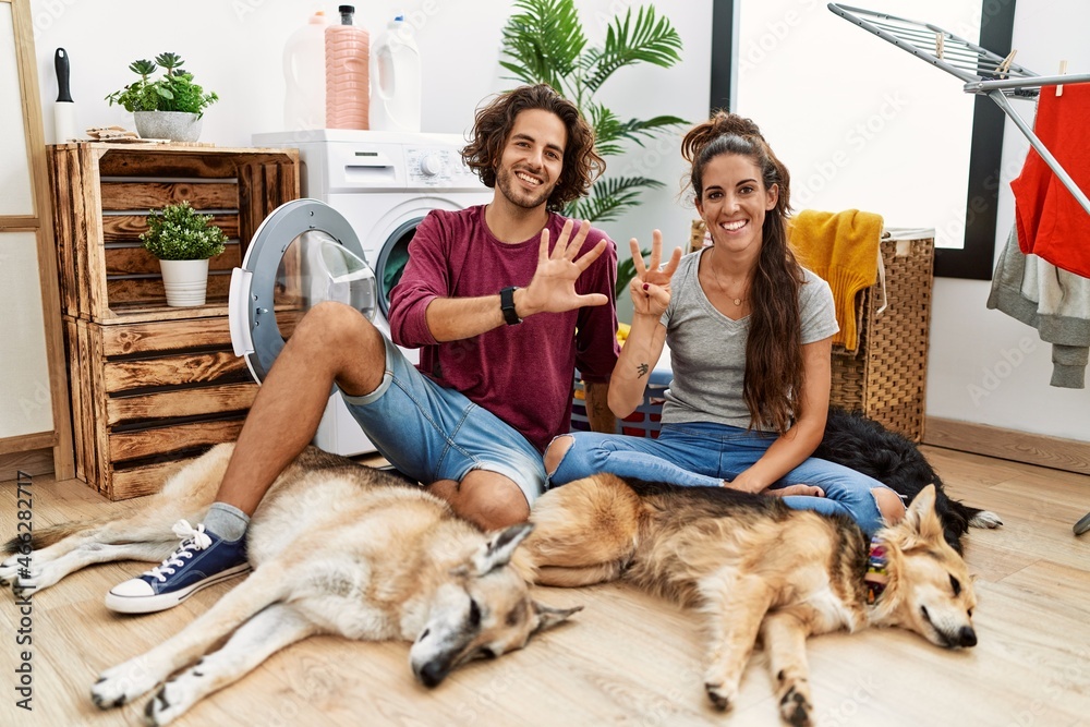 Young hispanic couple doing laundry with dogs showing and pointing up with fingers number eight while smiling confident and happy.