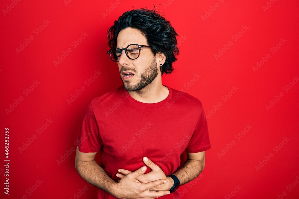 Handsome hispanic man wearing casual t shirt and glasses with hand on stomach because nausea, painful disease feeling unwell. ache concept. Handsome hispanic man wearing casual t shirt and glasses with hand on stomach because nausea, painful disease feeling unwell. ache concept.