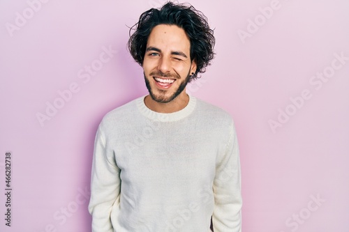 Handsome hispanic man wearing casual white sweater winking looking at the camera with sexy expression, cheerful and happy face.